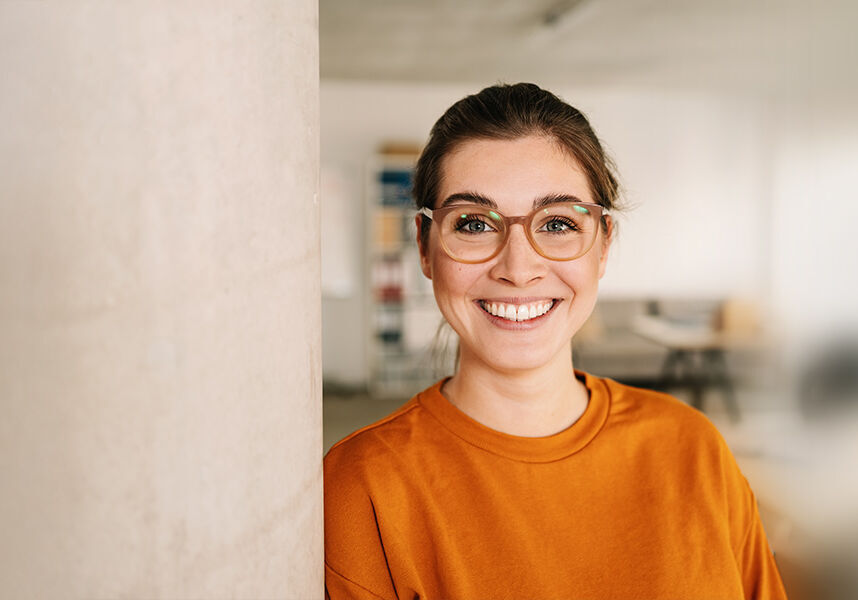 eine Frau in einem orangefarbenen Hemd und einer Brille, die an eine Wand gelehnt ist