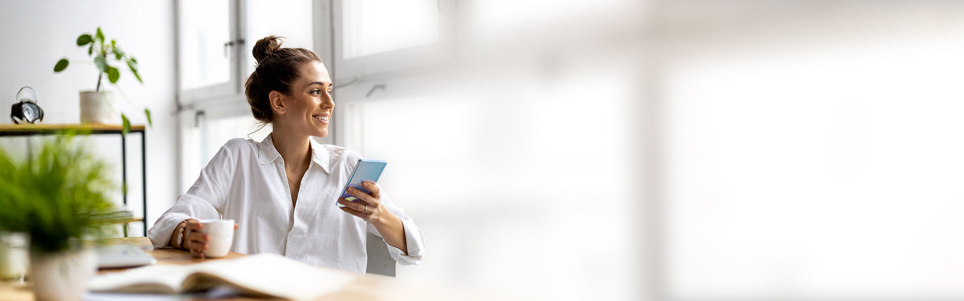 Junge Frau im Büro trinkt Kaffee und schaut auf ihr Smartphone.