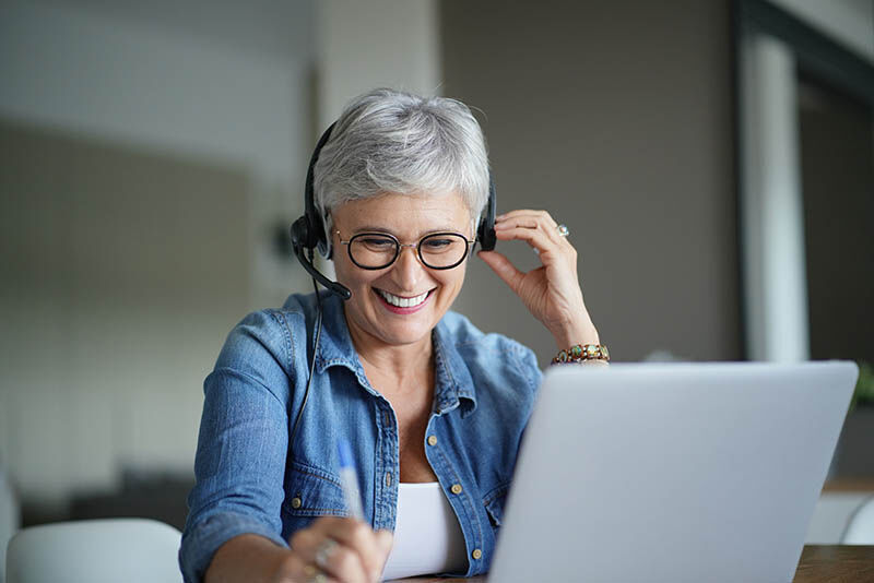 Frau mit kurzen grauen Haaren über 50 sicht an einem Laptop und Telefoniert mit einem Headset. Dabei Lacht sie freundlich