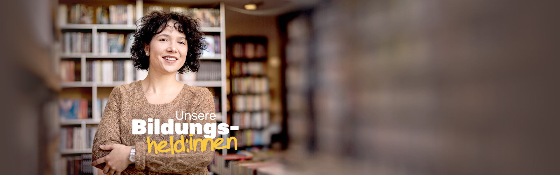 Website-Headerbild zum Thema "40 Jahre IBB - Bildungsheld:innen": Eine lächelnde gelockte Frau steht vor Bücherregalen.