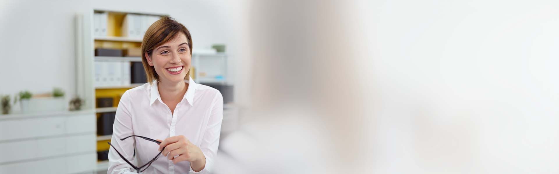 Lächelnde Businessfrau sitzt im Büro und hält ihre Brille in der einen Hand.